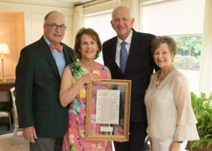 Four formally dressed older adults indoors, one woman proudly holding a framed award, all smiling after an academic achievement.