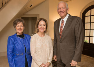 Three older adults in formal attire smiling together at the Guild Fall Coffee event indoors, enjoying an academic gathering.