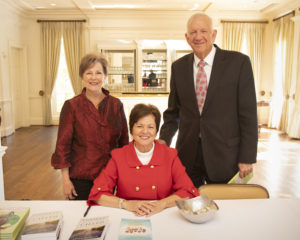 Three smiling adults seated at a table during a Christmas luncheon in a bright room with large windows and flowing curtains.