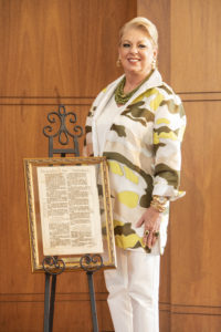 Smiling woman in patterned jacket stands by framed document on easel, indoors against wooden wall, during academic recognition event.