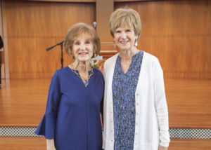 Two older women smiling together at an Installation of Officers event indoors, standing near a wooden stage and microphone.