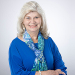 Jeanne Walker with long gray hair, blue top, and patterned scarf, posed as Guild President before a light backdrop.
