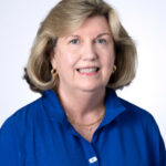 Guild President with blonde hair, royal blue blouse, and gold jewelry smiles in front of a light background.