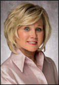Witham Jan Guild President with short blonde hair smiles in a light pink collared blouse, posing against a neutral gray indoor backdrop.