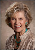 Barbara Boek with short blonde hair smiles in beige jacket and turquoise jewelry at Past Guild Presidents event indoors.