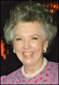 At an HCU ceremony, an Yvonne Cockrell with curly gray hair smiles in a pink floral dress and pearl earrings.