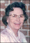 Ruth Coulson with glasses honors past Guild Presidents indoors, standing before a brick wall in a patterned blouse.