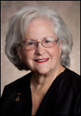 Marsha Eckermann with white hair and glasses smiles in front of a neutral backdrop, recognized for leadership among Guild Past Presidents.
