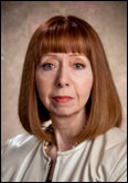 Peg Harman with straight red hair and bangs in a light jacket poses indoors during a formal Past Presidents portrait session.