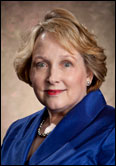 Kathryn Mayfield with short blonde hair in a royal blue suit jacket stands before a brown backdrop, honoring past Guild leadership.