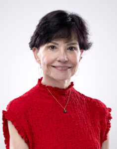 The HCU Guild President, a middle-aged woman with dark hair, wears a red high-neck top and a gold pendant necklace.