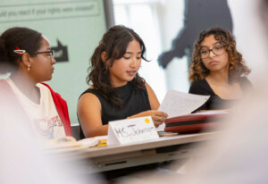 Students gather around a table in an academic setting as the center student reads aloud from an Honors College paper.