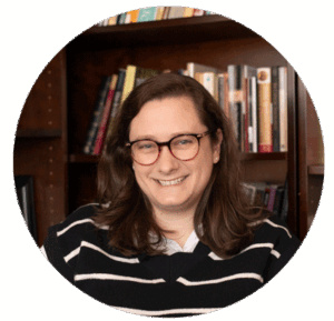Professor with long brown hair and glasses smiling, seated in front of a bookshelf lined with academic texts in an office.