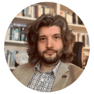 Professor with long, wavy hair and beard in checked shirt and blazer, seated in office with bookshelves in academic setting.