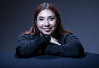 Student with long brown hair and vibrant blue eye makeup, smiling as she leans on a black table in a dimly lit classroom.