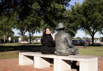 HCU Kinesiology grad wearing black sits smiling on a bench beside statue of man in hat, enjoying sunshine on HCU campus.