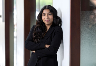 Student with long dark hair in a black suit stands confidently with arms crossed in an academic hallway, conveying connection.