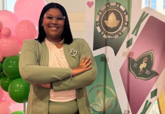Business student in a green suit smiles with arms crossed, standing by casino-themed décor and pink balloons at campus event.