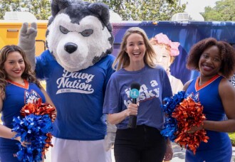 Husky mascot, reporter, and HCU cheerleaders smiling near a vibrant blood drive bus on the HCU campus.